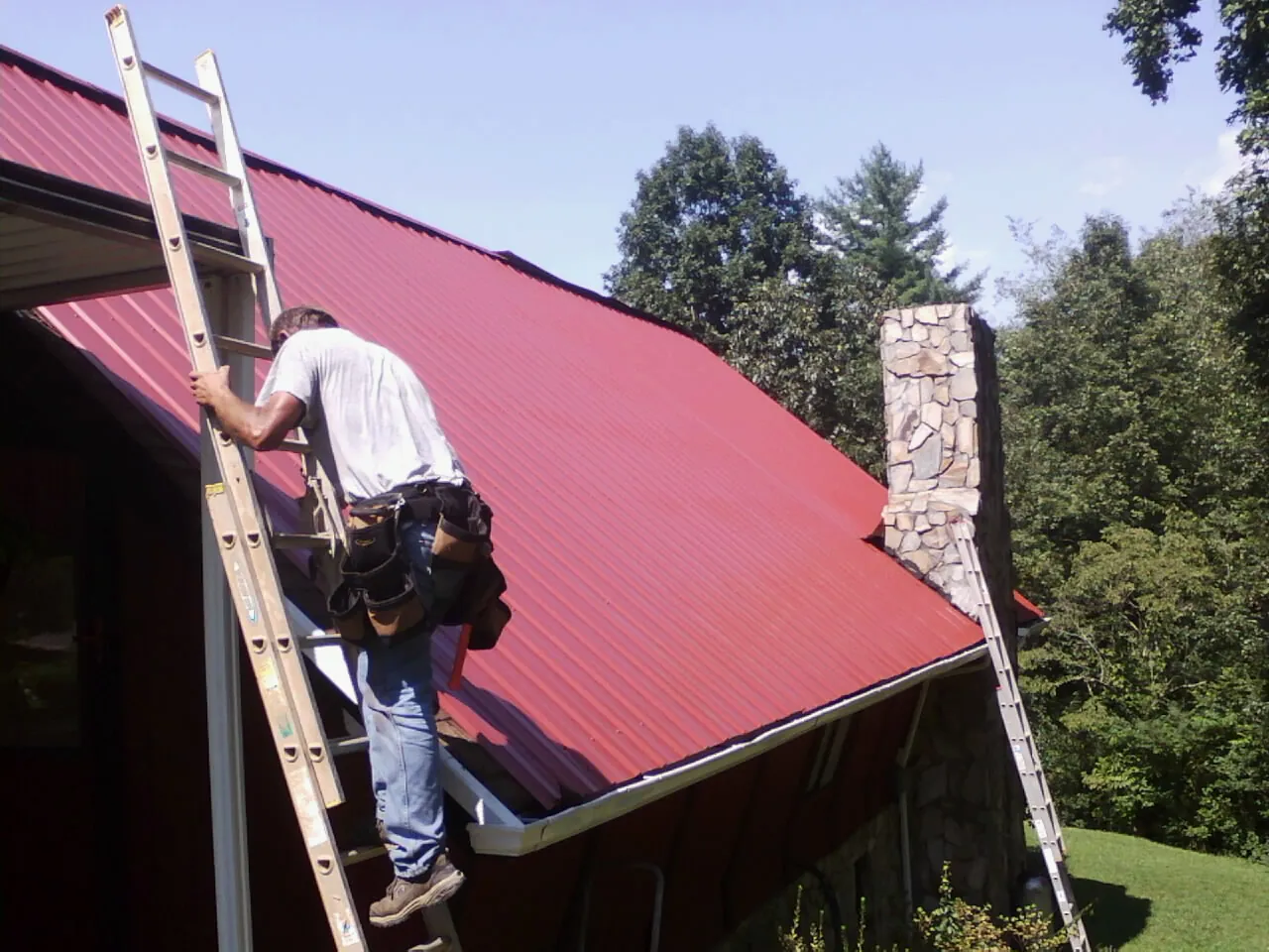 Roofer on a ladder beside a red metal roof and stone chimney — Soffit & Fascia Repair by Roofing Brothers in Glen Ellyn